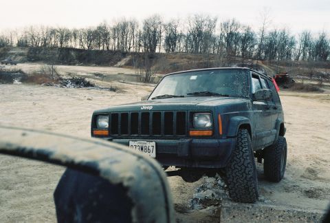 1995 Ford Bronco - A Jeep Cherokee XJ is parked on a dirt lot with trees in the background.