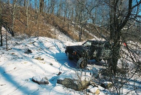 1995 Ford Bronco - A black Jeep Cherokee XJ navigates a snowy, wooded path with its driver's door ajar.