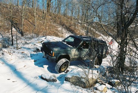 1995 Ford Bronco - A Jeep Cherokee XJ navigates a snowy, wooded terrain with one wheel lifted over rocks.