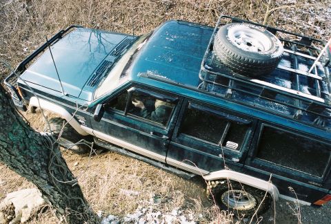 1995 Ford Bronco - This dark green SUV, equipped with a roof rack and spare tire, is shown off-roading through a wooded area with patchy snow.