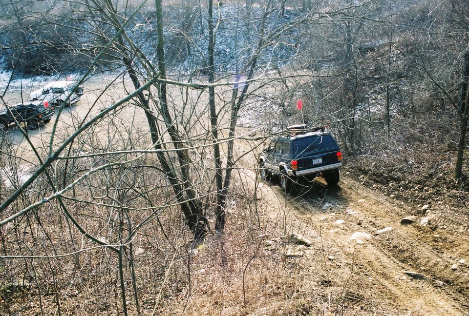 Jeep Cherokee XJ climbing a muddy trail, another vehicle behind it on a paved road