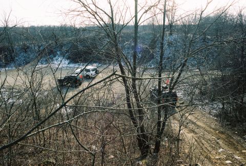1995 Ford Bronco - Two SUVs navigate a muddy track in a wooded area with patches of snow.