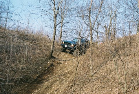 1995 Ford Bronco - A dark green Jeep Cherokee navigates a challenging off-road trail through a wooded area.
