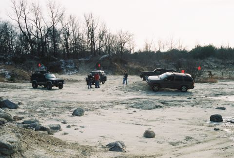 1995 Ford Bronco - Off-road vehicles are gathered in a sandy, outdoor area with bare trees.