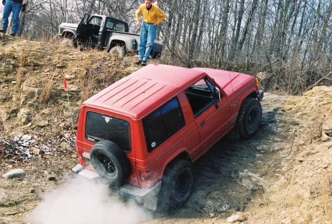 1995 Ford Bronco - A red off-road SUV navigates a challenging rocky terrain, with a pickup truck visible on the hill above.
