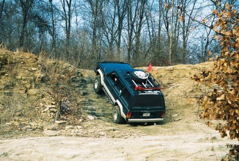 1995 Ford Bronco - A dark blue SUV navigates an off-road trail with a spare tire mounted on its roof rack.