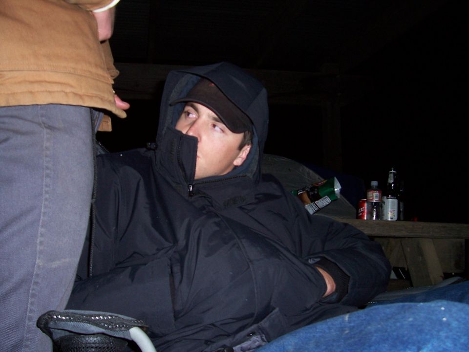Man in dark hooded jacket and black cap reclining at night near a picnic table with drinks