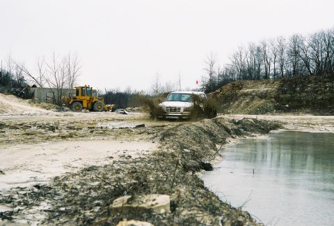 1995 Ford Bronco - A white SUV navigates a muddy obstacle course, with a yellow loader visible in the distance.