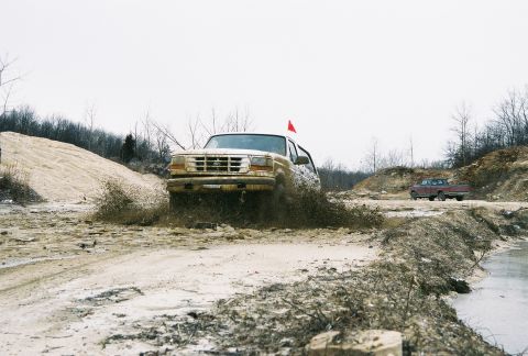 1995 Ford Bronco - A tan Ford Bronco navigates a muddy terrain, with another truck visible in the background.