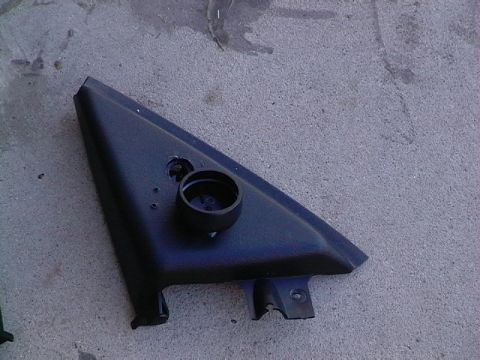 1999 Ford Taurus - A detached black plastic car part, likely a mirror housing trim, rests on a rough concrete surface.