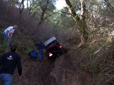 1998 Ford Ranger - A group of people attempt to free a Jeep Wrangler that is stuck in a steep, muddy incline surrounded by trees.