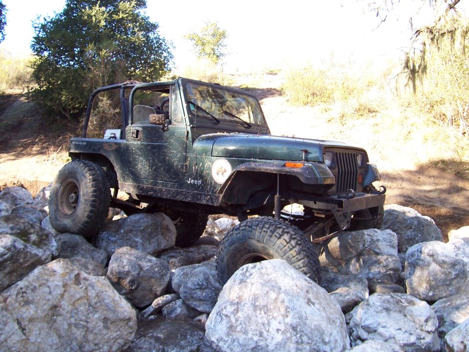 Green Jeep Wrangler on rocks, 'Jeep' badge visible on fender. Watermark says SUPERMOTORS