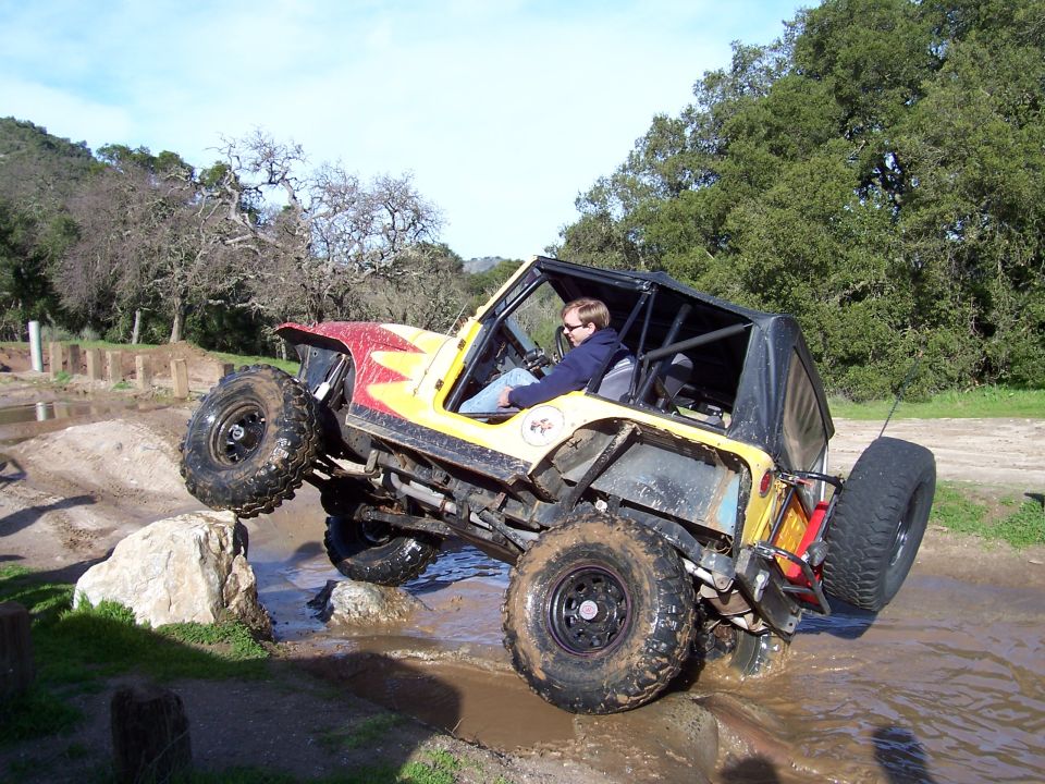 Yellow and red off-road jeep with roll cage, lifted suspension, and large tires, navigating a muddy obstacle course