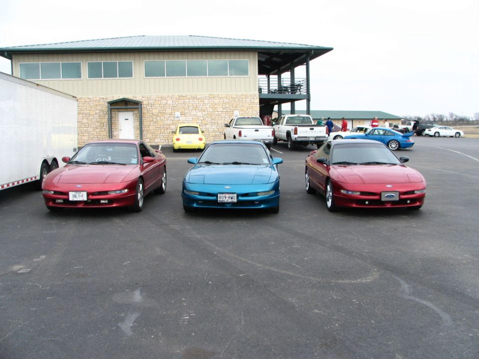 Two red Ford Probes and one blue Probe parked front-facing in a lot with a building and other cars behind