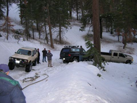 1999 Toyota Tacoma - Multiple vehicles are stuck in deep snow in a wooded area, with people trying to help.