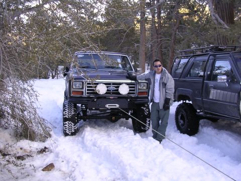 1999 Toyota Tacoma - Two lifted off-road vehicles are stuck in the snow with a man standing by the front vehicle.