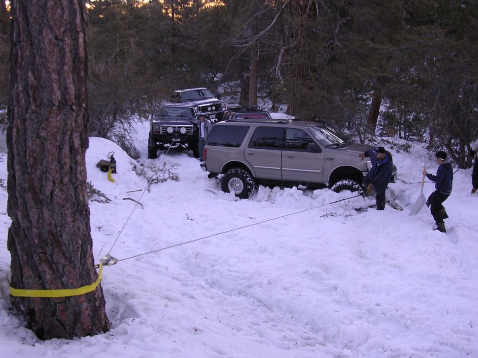 Several off-road vehicles are stuck in deep snow, with people attempting to free one with a winch and shovels