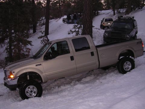 1999 Toyota Tacoma - A lifted tan pickup truck navigates a snowy incline, with other vehicles visible in the background.