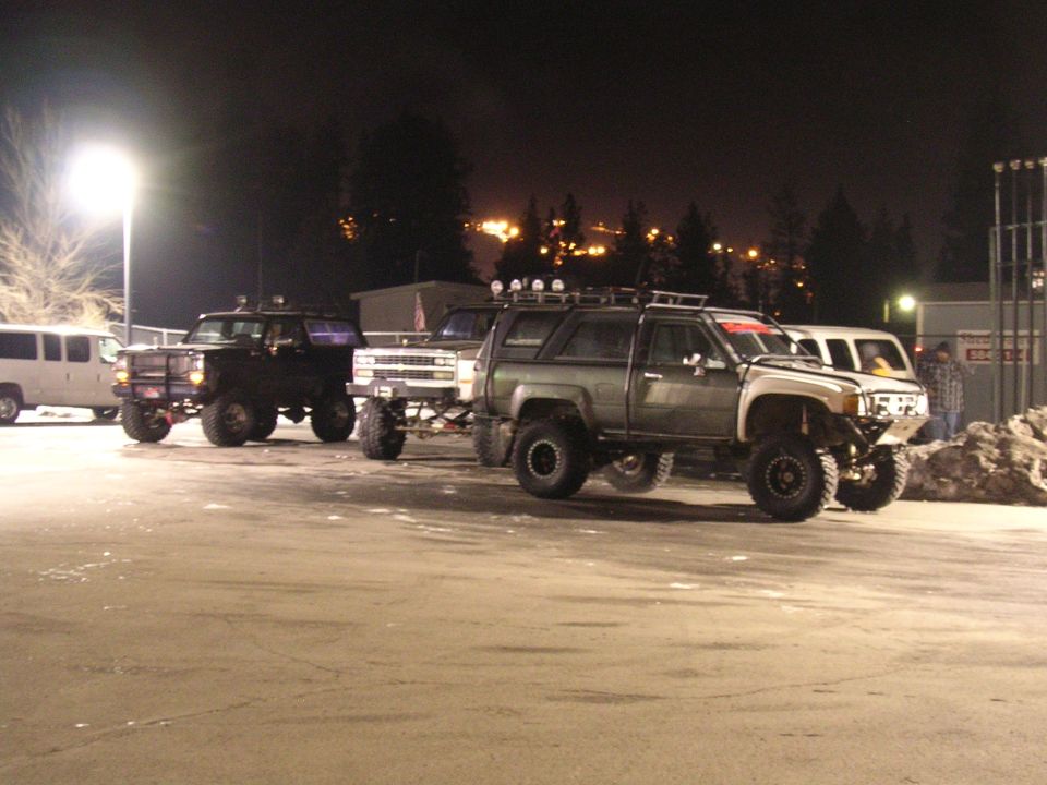 Several modified pickup trucks and SUVs parked at night, some with roof racks and off-road lights