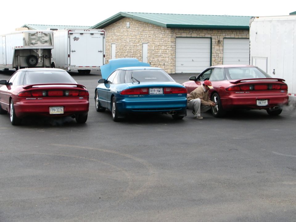 Three 1994 Ford Probes parked outside a building, one with hood up, one with a person working on a wheel