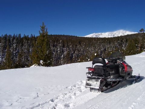 1994 Ford Bronco - Snowmobile rests in a snowy mountain landscape with a dense pine forest.