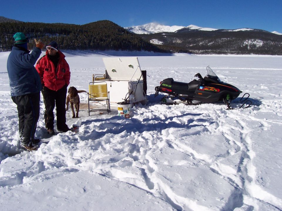 Two people and a dog stand near an ice fishing shelter and an Arctic Cat snowmobile on a frozen lake with mountains in the background