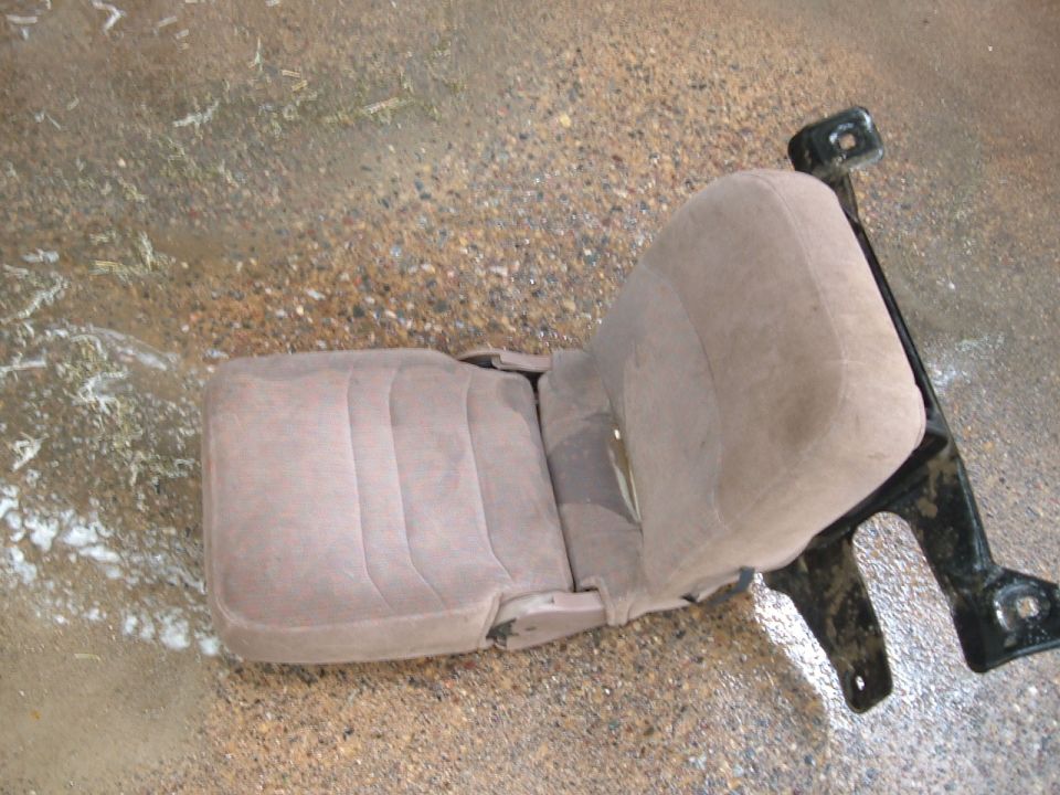 Tan fabric car seat with black metal mounting brackets, on a wet, gravelly surface