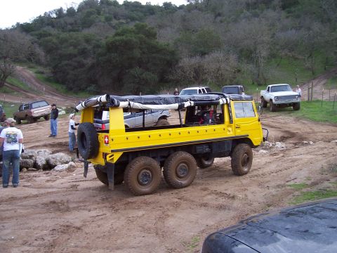 1998 Ford Ranger - A bright yellow Pinzgauer 6x6 all-terrain vehicle with a rolled-up soft top and rear-mounted spare tire, parked on a muddy off-road area. S…
