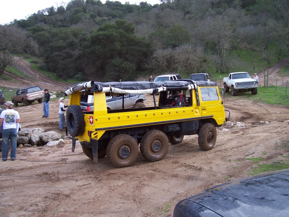 Yellow Pinzgauer 6x6 off-road vehicle parked on muddy trail with trucks behind