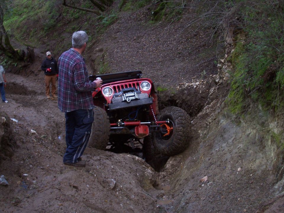 Red Jeep navigating a steep, muddy ditch on a trail. Guy in plaid shirt watching