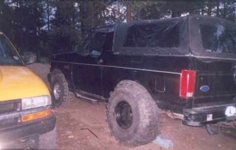 1995 Ford Bronco - A black Ford Bronco with a soft top and large tires is parked next to a yellow truck.