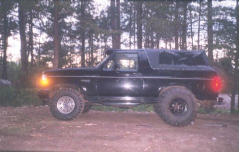 1995 Ford Bronco - Side view of a black Ford Bronco with a soft top, parked outdoors in a wooded area.