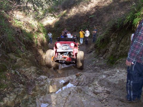 1998 Ford Ranger - A red off-road vehicle tackles a challenging rocky and muddy terrain, with spectators observing the action.