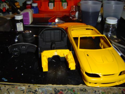 1979 Ford Bronco - Model car parts, including a yellow body and black interior, are laid out on a workbench next to various hobby supplies.