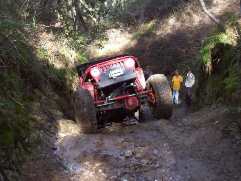 1998 Ford Ranger - A modified red jeep navigates a challenging muddy trail with spectators observing the difficult climb.