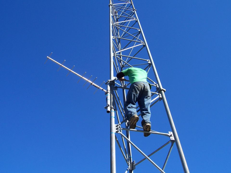 guy climbing a tall metal tower with antennas against a blue sky