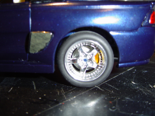 Blue model car rear wheel with chrome five-spoke rim and yellow brake caliper. Textured vent on fender