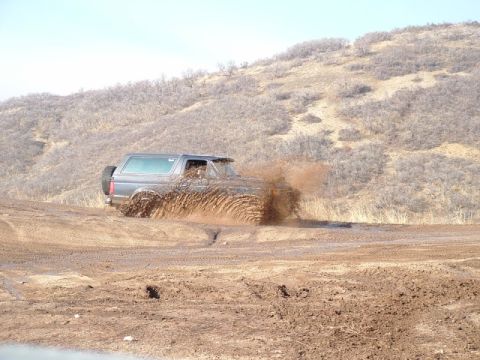 1991 Ford Bronco - A gray Ford Bronco navigates a muddy off-road course, sending dirt and water flying.