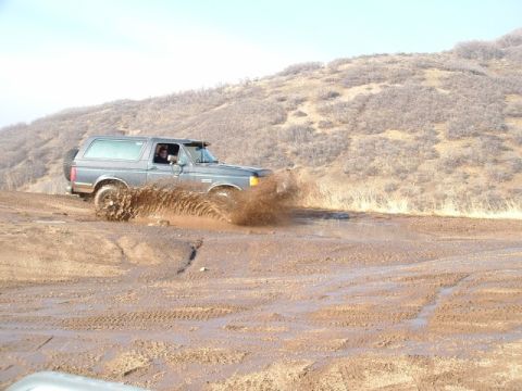 1991 Ford Bronco - A gray 1991 Ford Bronco navigates a muddy off-road trail, kicking up a large spray of water.