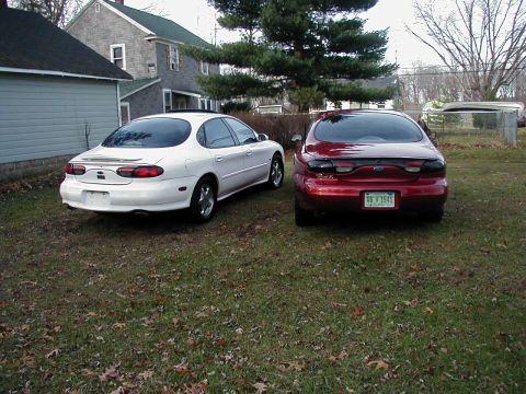 1997 Ford Taurus - Two sedans, one red and one white, are parked in a grassy yard with houses in the background.