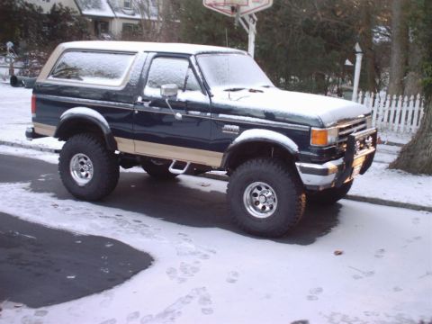 1989 Ford Bronco - A lifted Ford Bronco sits on a snow-covered road, its dark blue paint contrasting with the tan trim and the wintery surroundings.