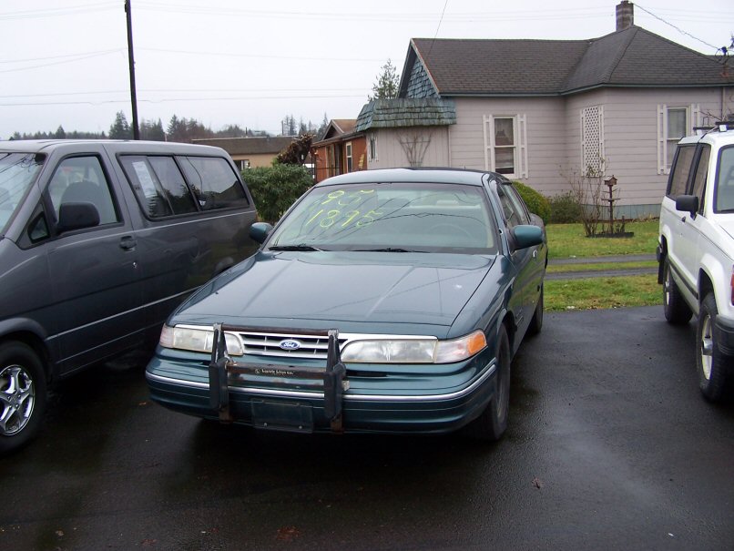 Dark green Ford Crown Victoria sedan with front push bar and yellow windshield writing, parked on wet lot