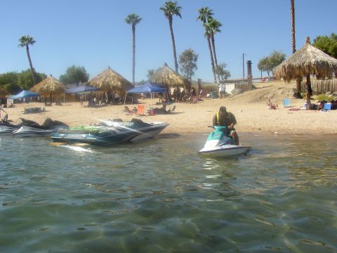 1990 Ford Bronco - A person rides a jet ski in the water near a sandy beach with palm trees and thatched huts.