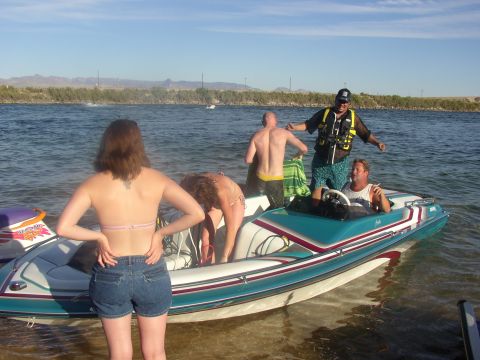 1990 Ford Bronco - People stand in and around a white-and-teal ski boat at the edge of a lake, with a woman in shorts watching from shore.