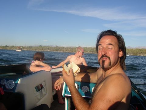 1990 Ford Bronco - man with a goatee makes a face in a boat on a lake while two other passengers sit toward the bow under a blue sky.