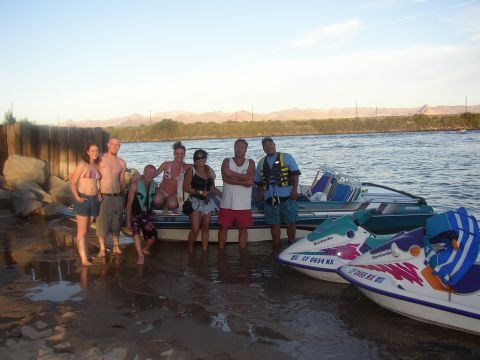 1990 Ford Bronco - group of seven poses on and beside a beached ski boat and jet skis at a lakeshore with mountains glowing at dusk.