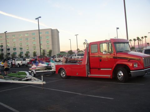 1990 Ford Bronco - red Freightliner ramp-bed truck lettered "Easy Rider" sits in a parking lot beside a boat on a trailer at dusk.