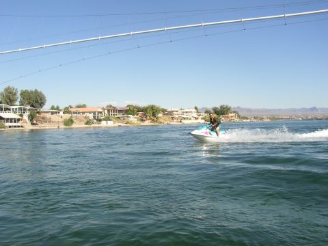 1990 Ford Bronco - rider on a teal-and-white jet ski crosses a wide river under a low suspended cable, with hillside homes on the far bank.