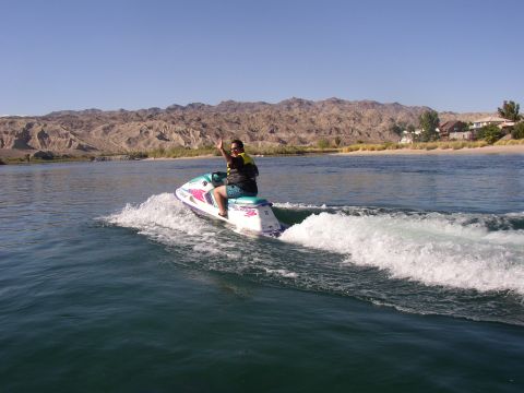 1990 Ford Bronco - person rides a teal-and-pink jet ski across river water, throwing spray, with rugged desert mountains behind.