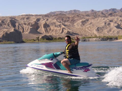 1990 Ford Bronco - man in a yellow life vest waves while riding a teal-and-pink Sea-Doo personal watercraft on a river below desert hills.